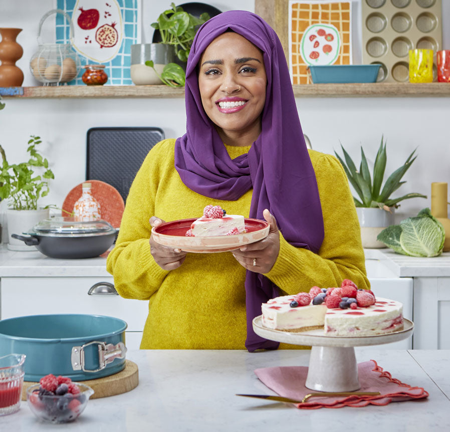 Nadiya smiles at the camera, holding a plate with a slice of Raspberry Ripple cheesecake