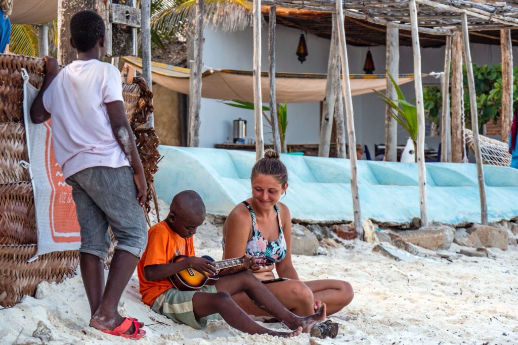 Reisende sitzt mit zwei einheimischen Kindern am Strand und schaut ihnen beim Spielen zu