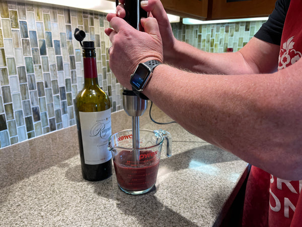 Photograph showing using an immersion blender to aerate a Cabernet Sauvignon, with a bottle in the background.