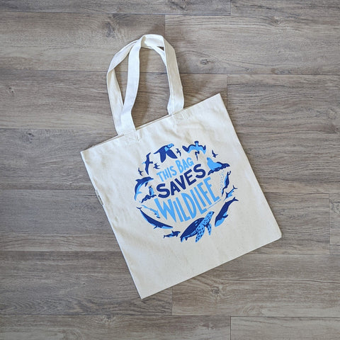 An organic cotton tote bag with the words "This Bag Saves Wildlife" and illustrations of ocean wildlife screen printed on it in two shades of blue