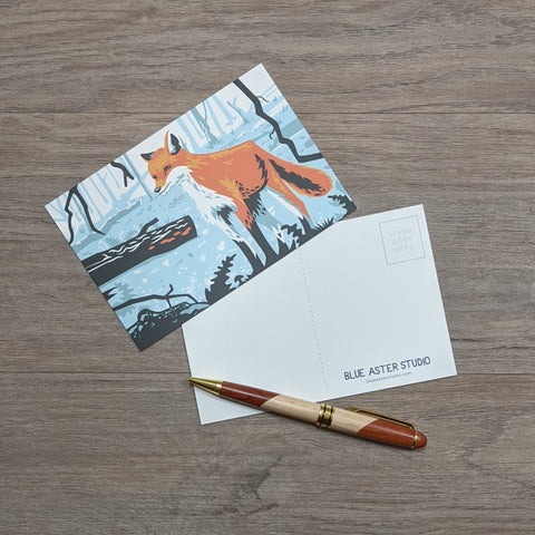 A postcard featuring an illustration of a red fox sitting next to a pen.