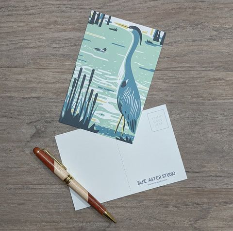 A postcard with an illustration of a great blue heron on it sitting next to a pen.