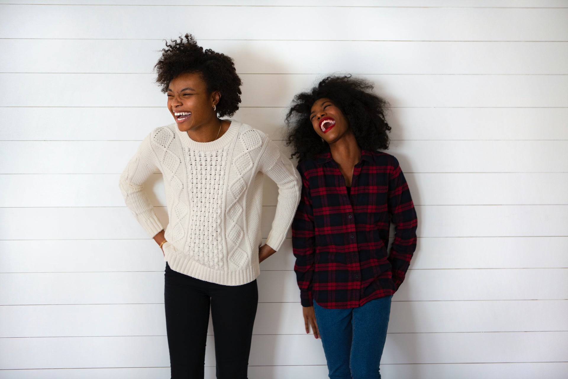 Two friends laugh in sweaters against a wall