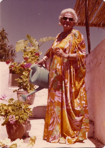 A smiling woman in sunglasses and a glorious yellow/pink/orange caftan is standing in the sun, watering the plants on her patio garden with a green watering can. The photo looks to be from the mid-seventies.