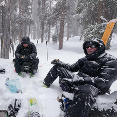 a stormy day, two guys sitting on their backpacks in a snow storm, with snow piling up on them