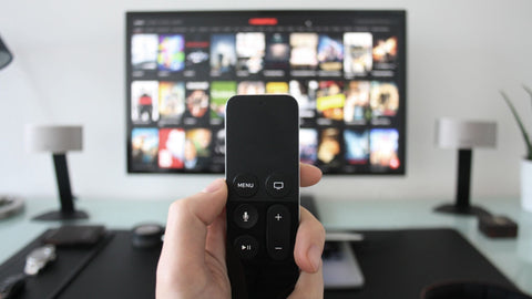 A hand holding up a small black remote control. Background is blurry with a TV screen, desk, and two small lamps.