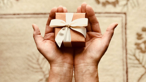 Two hands holding a small tan gift box with white ribbon and a bow