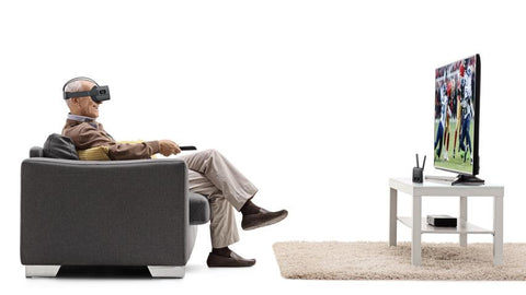 Elderly man wearing Vision Buddy headset sitting on a couch and watching TV that's on a table with a rug beneath the table