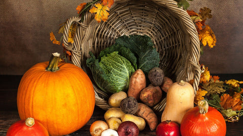 A cornucopia with orange and red pumplins, potatoes, and leafy green vegetables. It is decorated with orange, red, and yellow leaves. 