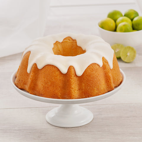 Image of key lime bundt cake with white icing sitting on a white cake stand with a bowl of limes in the background.