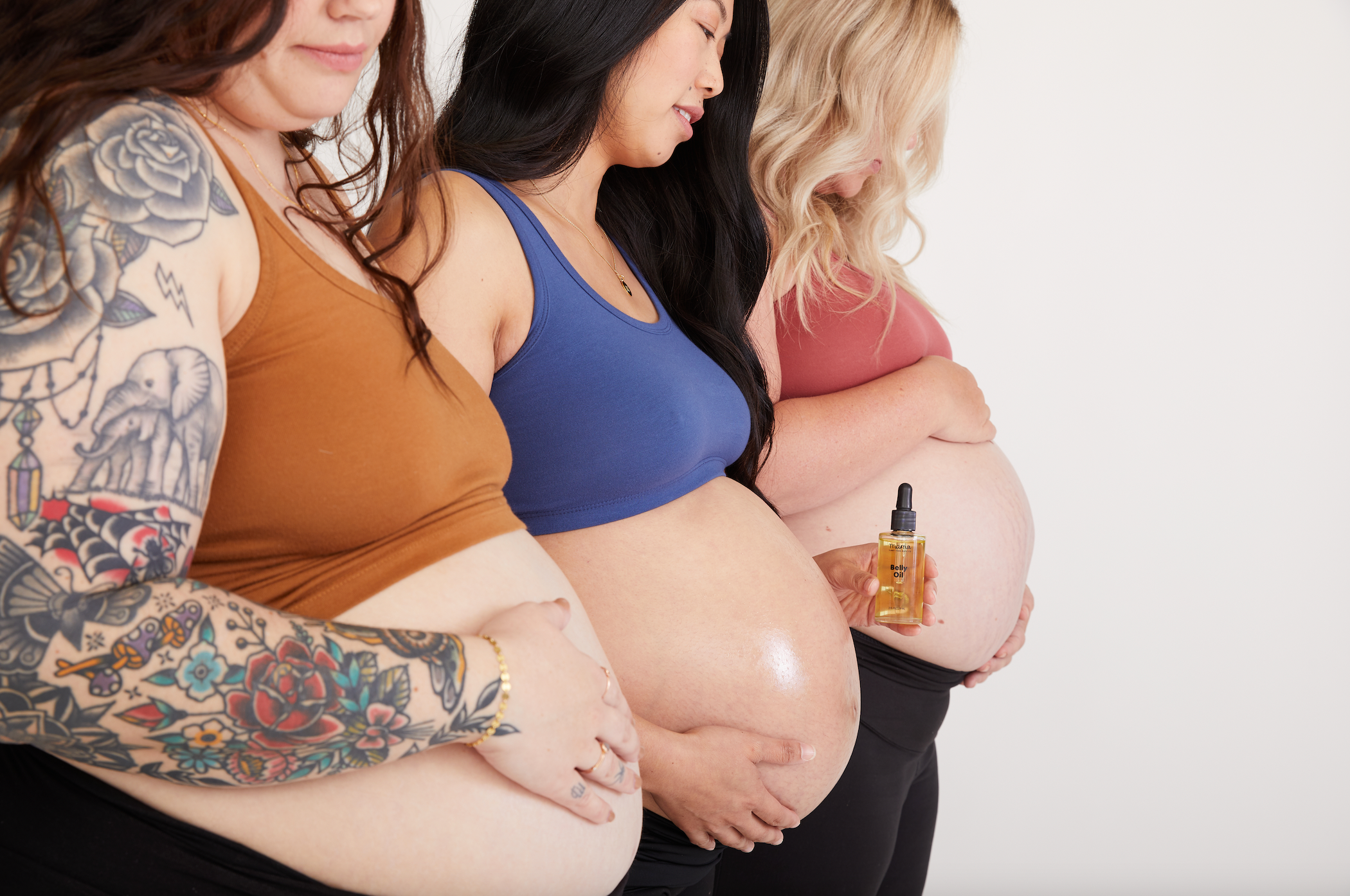 Three pregnant women, the woman in the middle is holding Belly Oil