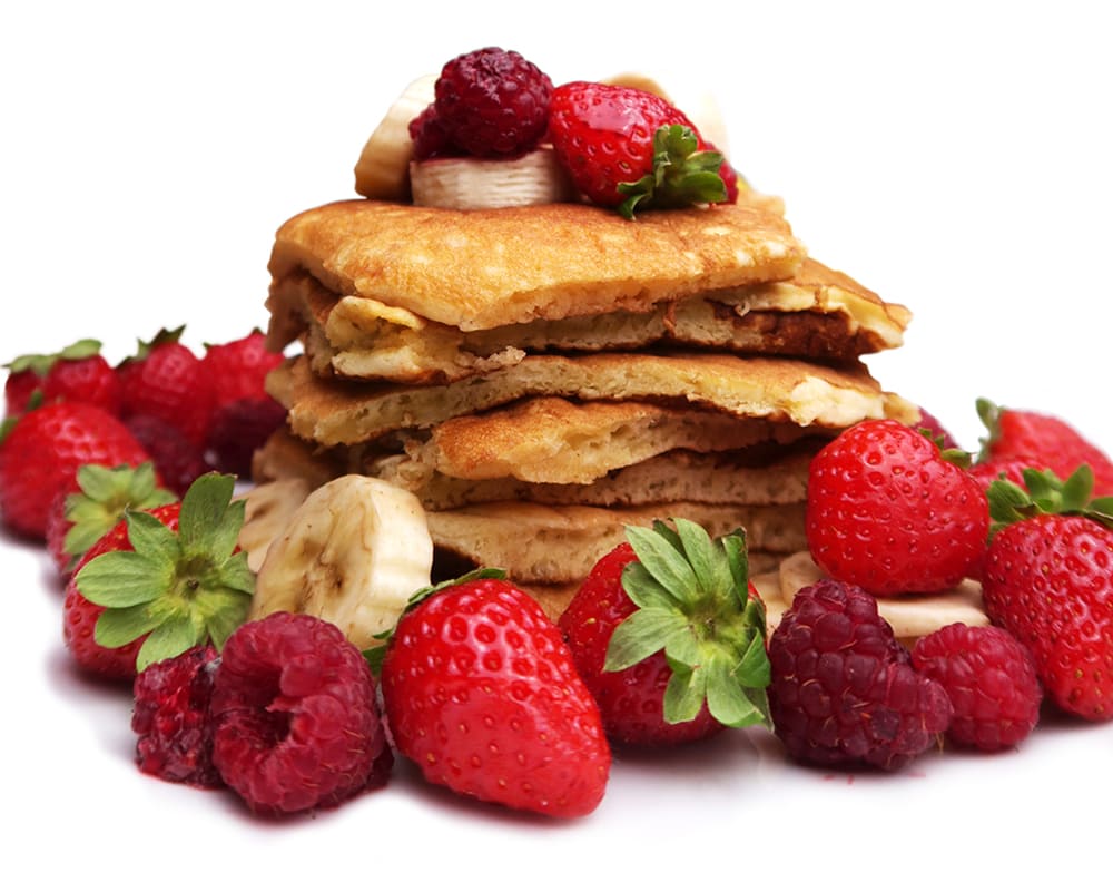 Pile de pancakes entourée de fraises et de framboises avec quelques morceaux de bananes par-dessus sur un fond blanc.