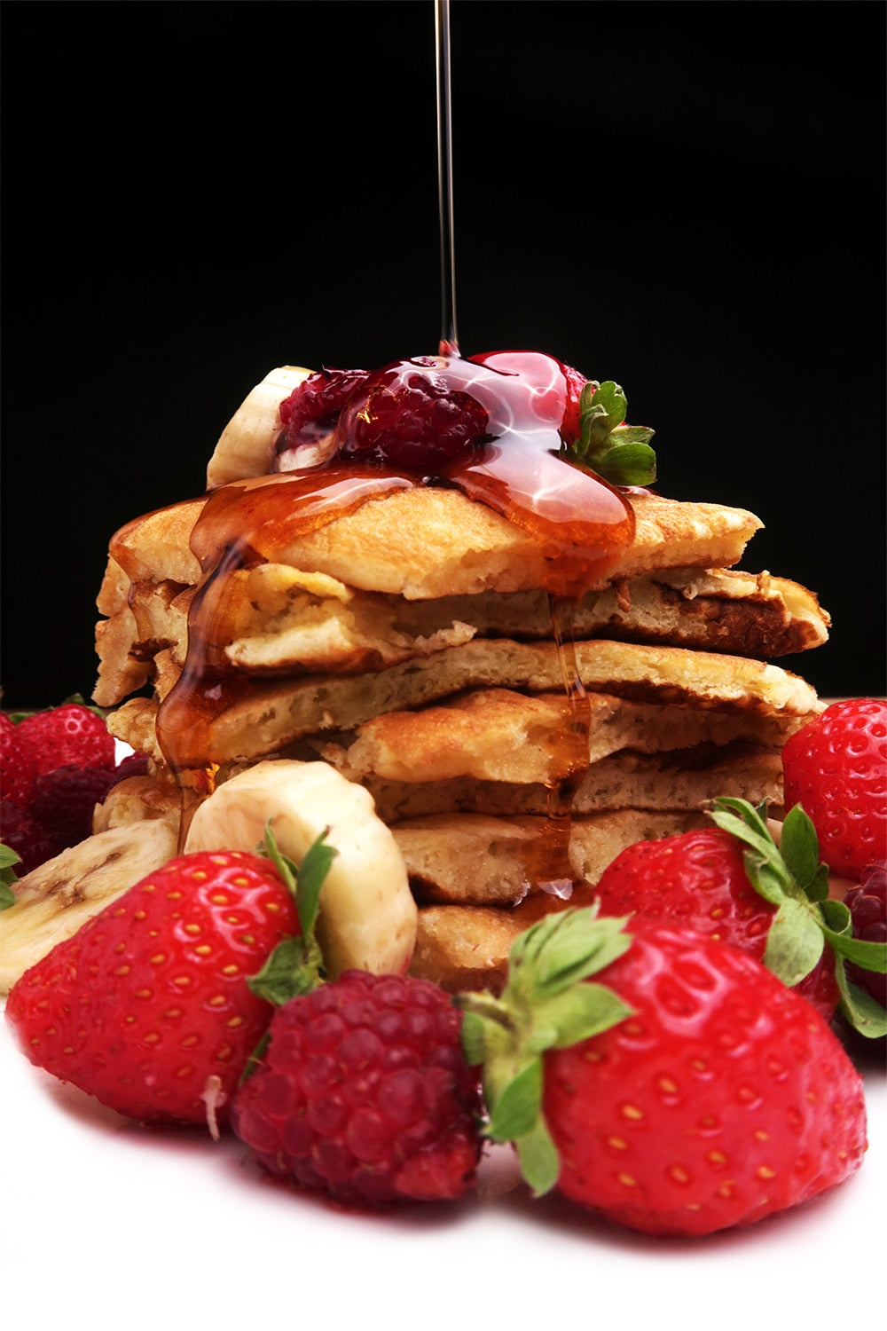 Pile de pancakes entourés de fruits rouges avec un filet de sirop d’érable versé par-dessus.