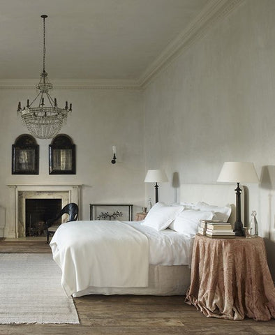 Large open concept bedroom with large bed on the right side. It has white linens on it, a large area rug, and a crystal chandelier up above.