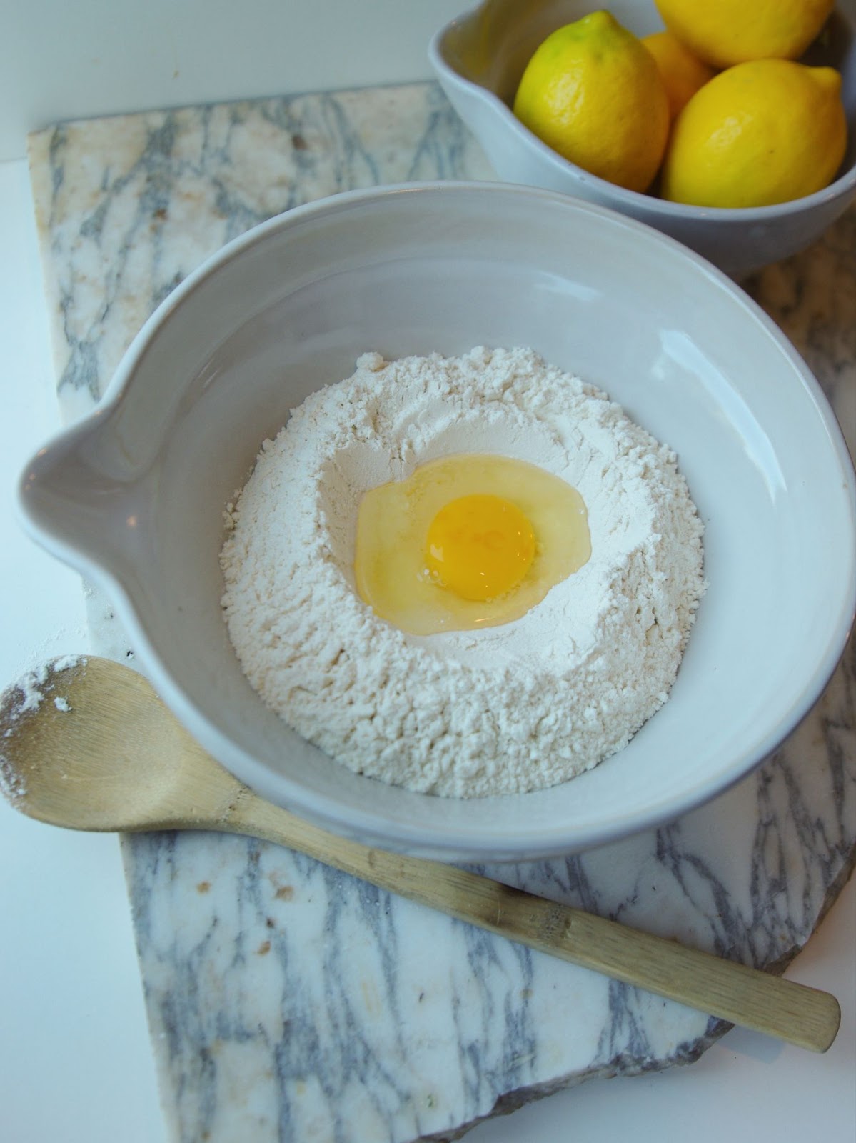 Egg and flour inside mixing bowl with spout next to wooden spoon and lemons.