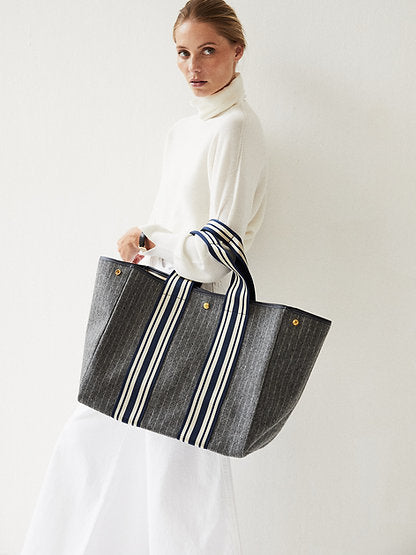 A woman all in white on a white background is carrying a grey striped wool tote and looking at the camera