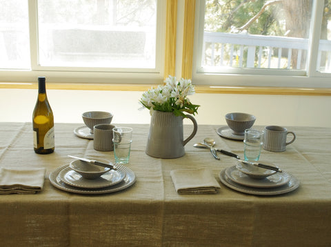 Kitchen table with grey plates, bowls, and vase. Bottle of wine sits on the table and yellow light streams through the window.