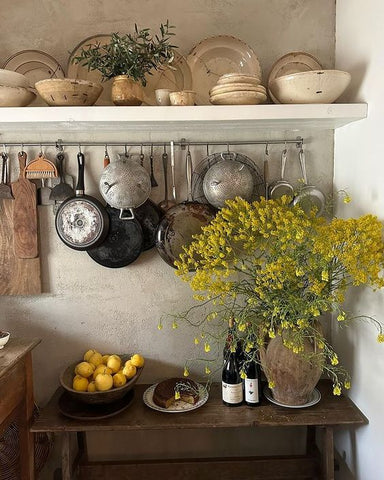 Rustic French kitchen shelf with pots has a big vase of yellow flowers below and a bowl of yellow lemons in a wooden vessel.