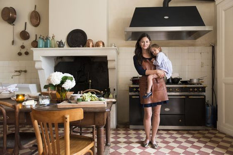 Mimi Thorisson stands with her son in her arms. She's in her kitchen wearing a short black dress. The kitchen floor has red and white checkered tiles and a large black range.