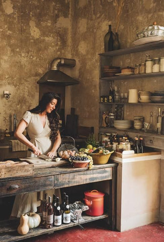 Woman stands at a sink in an old French kitchen cutting vegetables. She wears a white linen dress and has long dark hair