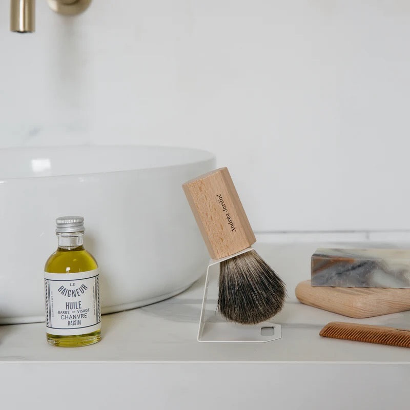 Shaving brush on stand next to glass jar of oil and bar of soap near sink.