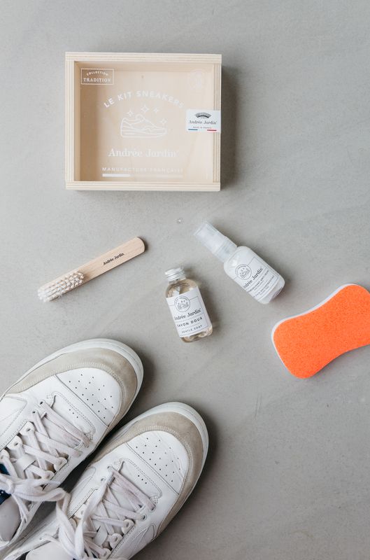 A pair of sneakers is on a grey background. Surrounding the sneakers are a wooden box, a small brush, two small bottles of shoe cleaner, and an orange sponge.