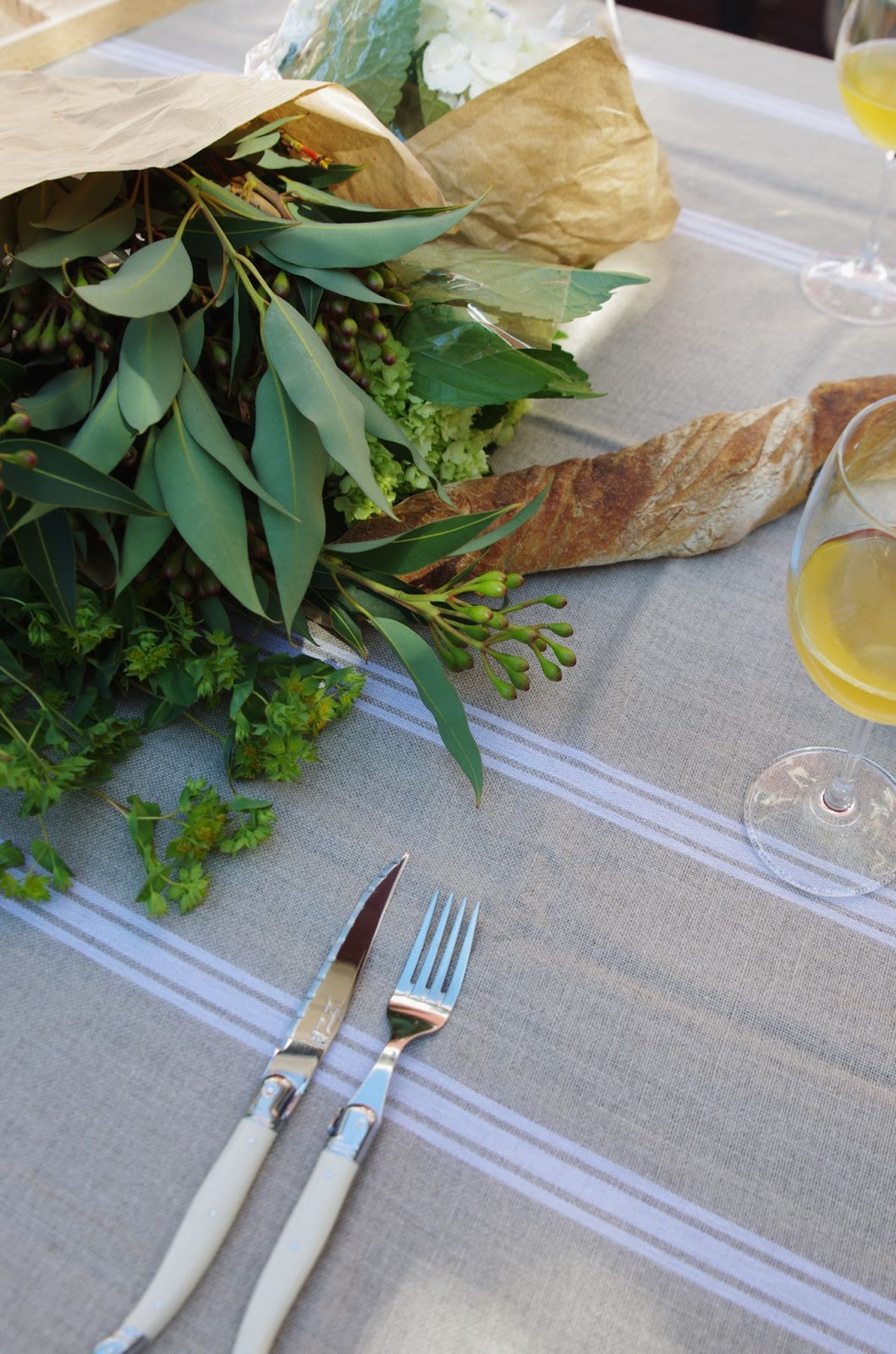 Outdoor dinner table setting with linen tablecloth, bottles and glasses of wine, bread, fruit, cutlery, napkins and flowers.