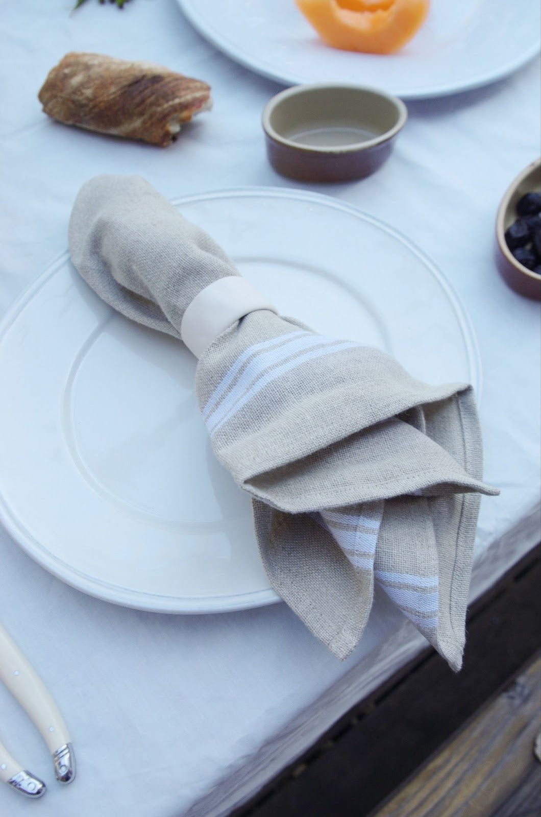Linen napkin with white stripes at hem on a plate next to bread, cutlery, and small bowls.