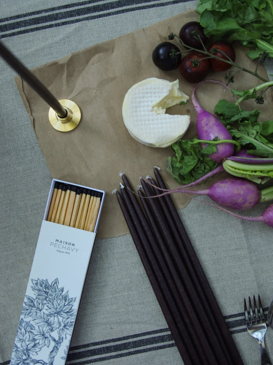 Black candles and box of long matches sit on a table. There is a piece of brie cheese and purple radishes in the frame.