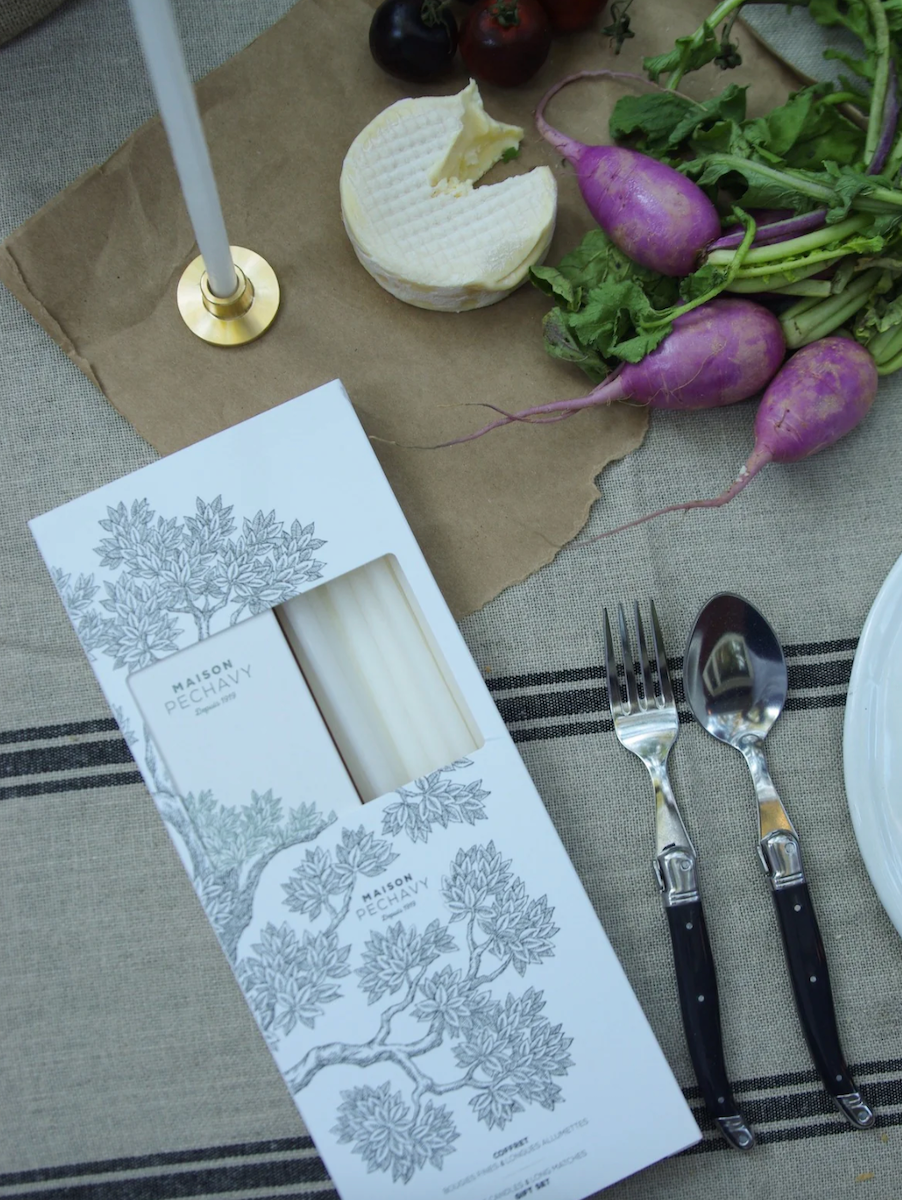 Package of white candles and long matches on table surrounded by radishes and laguiole cutlery