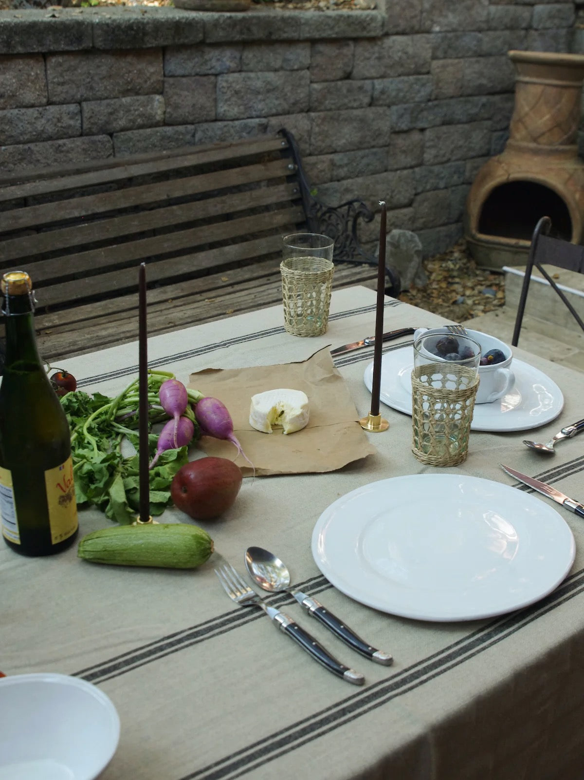 Black and natural striped linen tablecloth sits on a table outside. The table is set with white plates, black cutlery, black candles, and seagrass glasses.