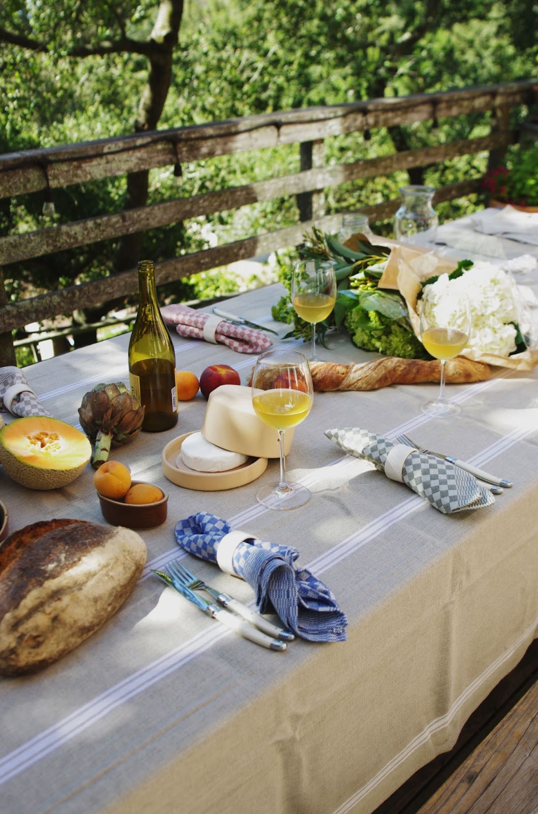 Outdoor dinner table setting with linen tablecloth, bottles and glasses of wine, bread, fruit, cutlery, napkins and flowers.