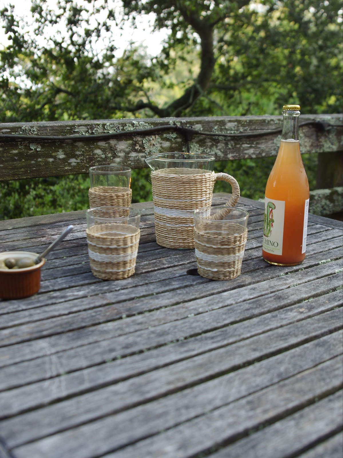 Outdoor table setting of seagrass wrapped glassware and pitcher next to bottle of wine and ramekin full of olives.