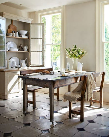 Wooden table sits in a brightly lit kitchen. Flowers are on the table and the pantry behind the table has it's doors open. The floor is white tile with small black tiles in between.