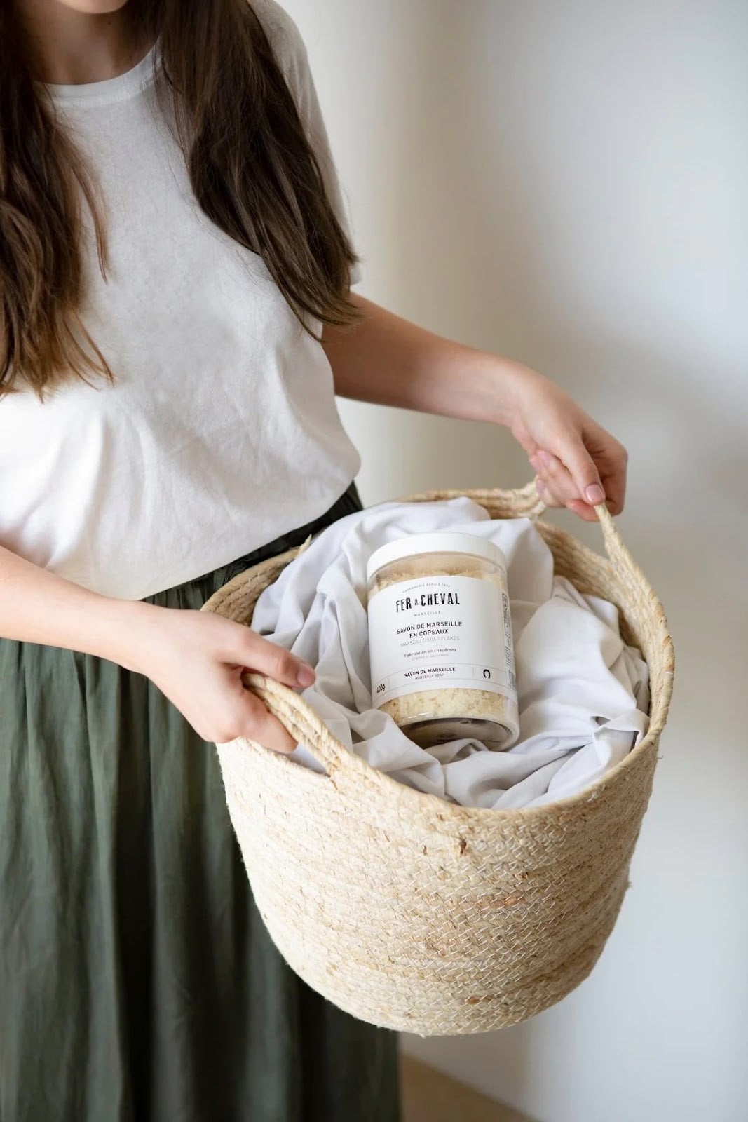 Person holding laundry basket holding linens and tub of Marseille soap flakes.