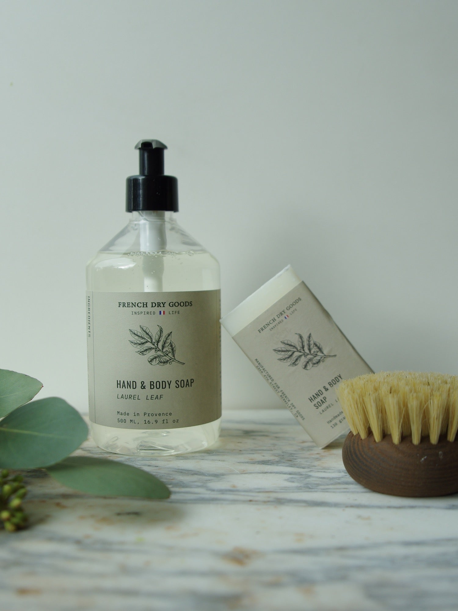 French Dry Goods Laurel Leaf Soap in liquid pump and bar soap on marble counter next to eucalyptus and body scrub brush.