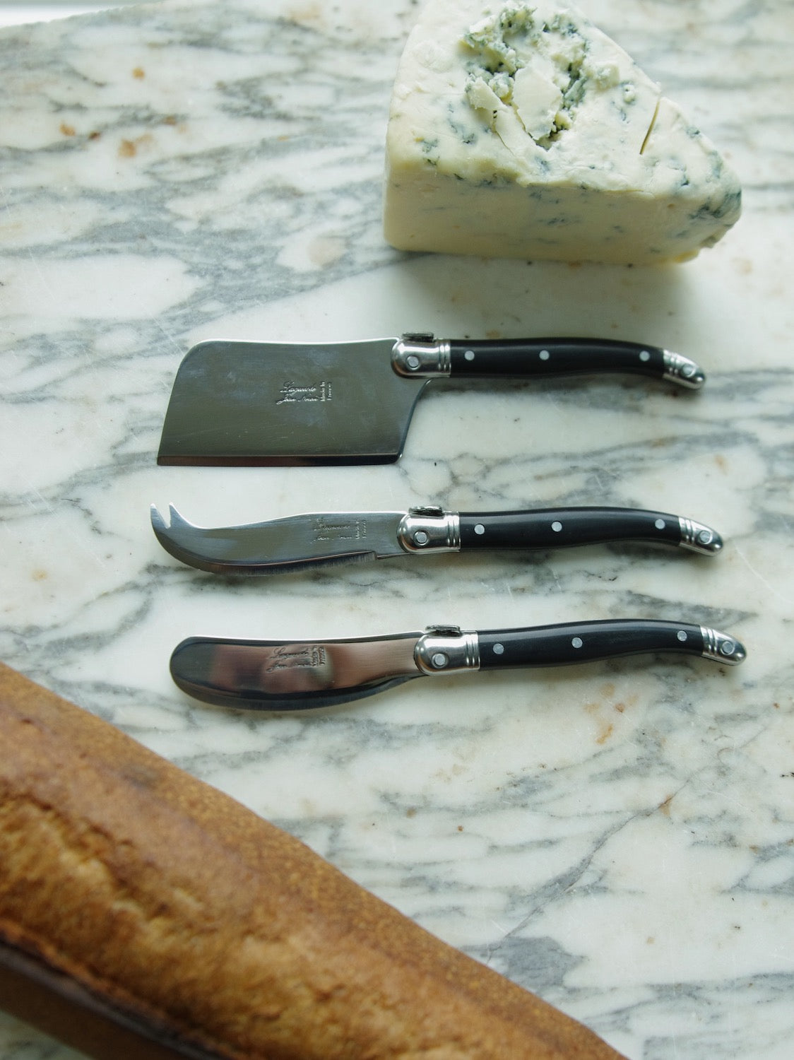 Laguiole mini cheese spreader, fork and knife with black handles next to block of cheese and baguette.