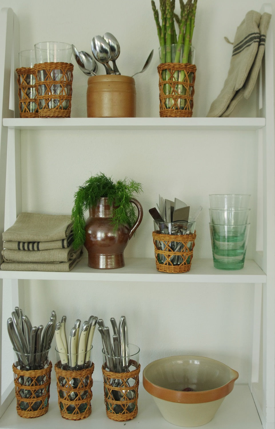 Kitchen shelves with linens, vintage French pottery, rattan wrapped cups holding Laguiole utensils, and clear glassware.