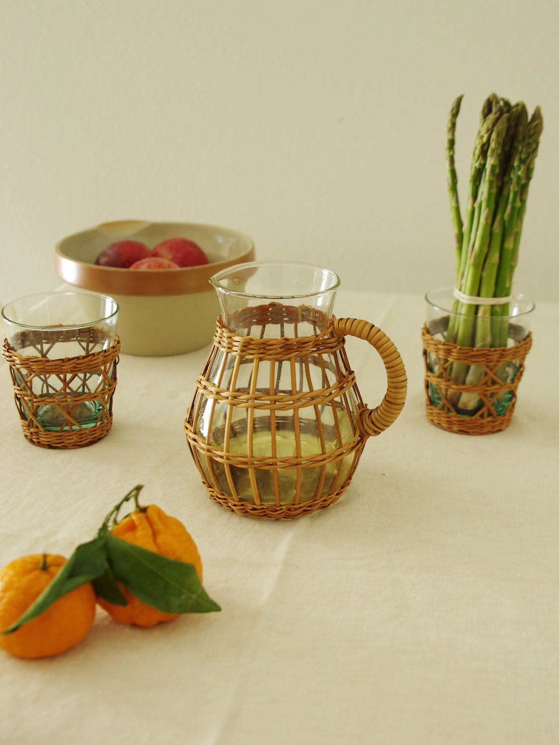 Rattan wrapped glass pitcher and tumblers with fruit and vegetables on table next to vintage French bowl.