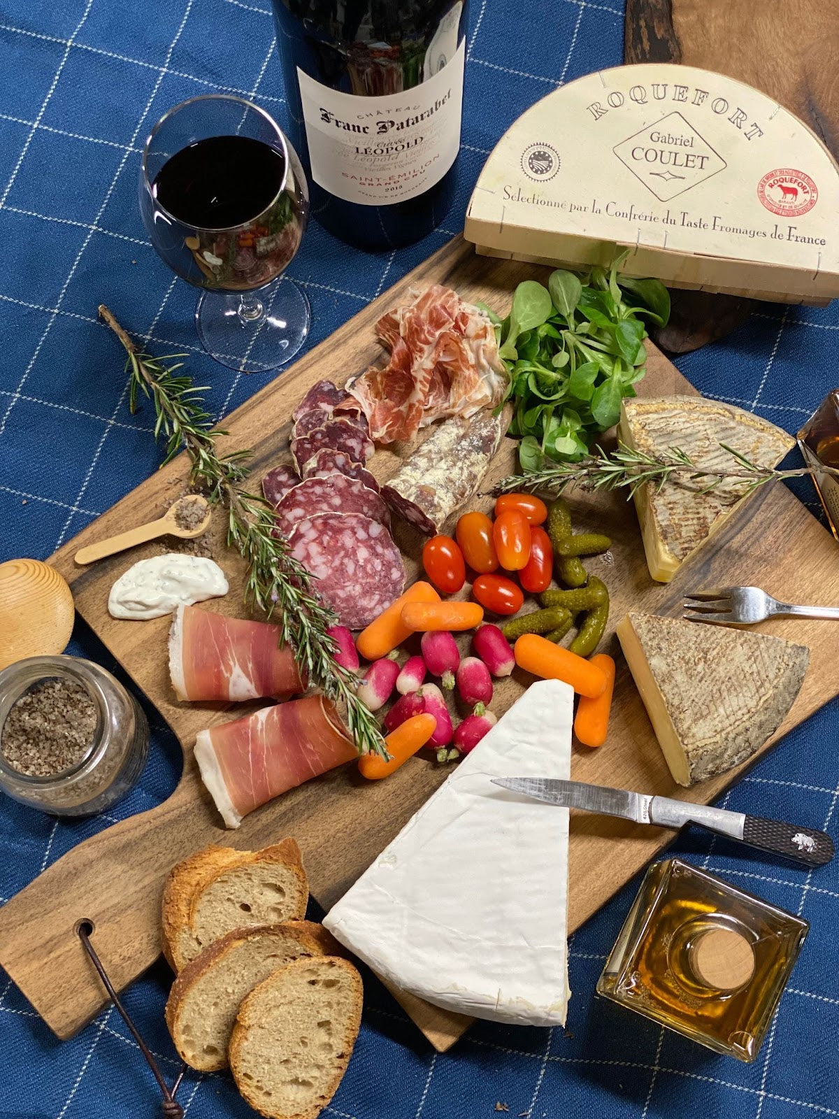 Wooden charcuterie board with veggies, cheeses, meat, herbs all on top. Next to bottle and glass of wine, cheese wedge, and a spread.