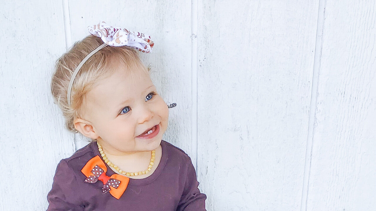 Baby Girl Wearing Baltic Amber Teething Necklace from Amber Guru in Butterscotch Color and White Background