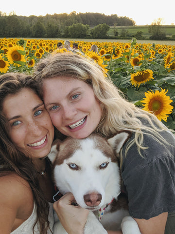 Emma with her fiance, hugging their dog in front of a field of sunflowers