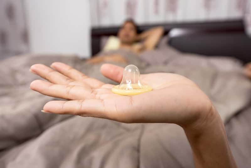 Woman's hand holding a delay condom with man in bed in background