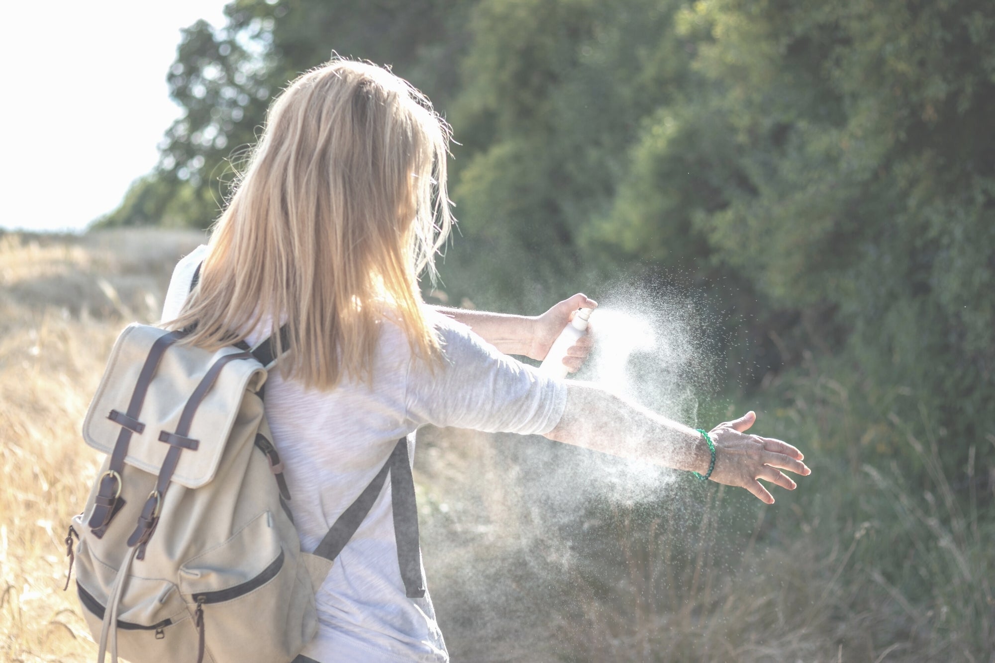 eine Frau mit Rucksack sprüht sich bei einem Spaziergang mit Insektenspray ein