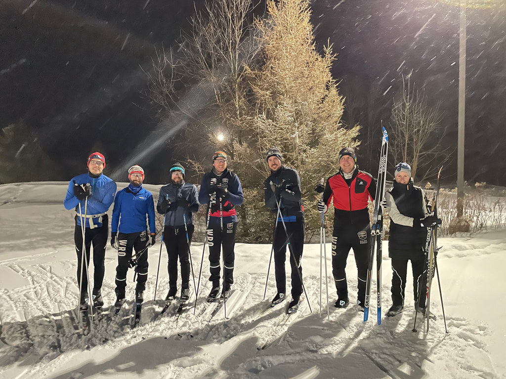 A group of cross-country skiers stands under a streetlamp at night