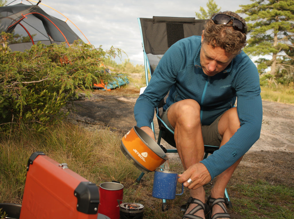 A man sitting in front of a camp stove pours water into a mug