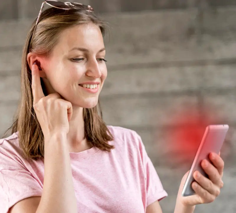 Woman smiling, using smartphone with wireless earbud, blurred background.