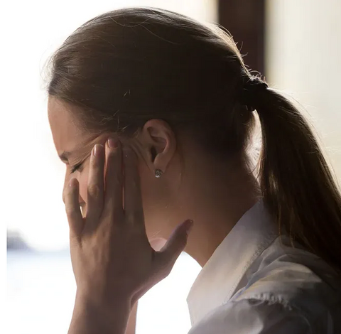 Woman with a pony tail, fingers to her temples indicating she has a headaches, eyes are closed.