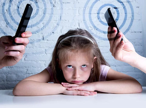 image of little girl with her chin on her folded hands on a table. Two hands are holding cell phones to the left and right of her head showing how much radiation is coming off of the phones into her head