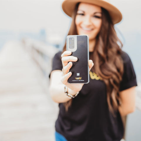 Image: a young woman holds a cell out as if she is taking a selfie. Her expression pleasant with a smile. This image illustrates the direct contact between a user and cell phone radio frequency radiation, emphasizing the focus on studies regarding its potential health effects discussed in the blog post.